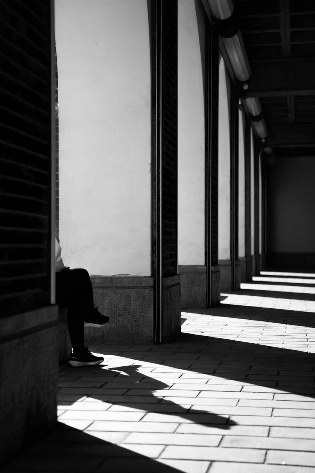 A solitary figure sits in shadow along a corridor of repeating columns and light, reflecting the isolating patterns of emotional and physical symptoms addressed in depression counseling.