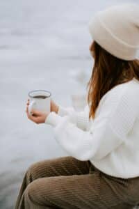 Woman holding a warm coffee cup outdoors, wearing a winter hat and sweate,r with snowy scenery behind her