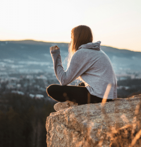 A woman sitting on a mountaintop overlooking a wide landscape, reflecting quietly while taking in a peaceful view