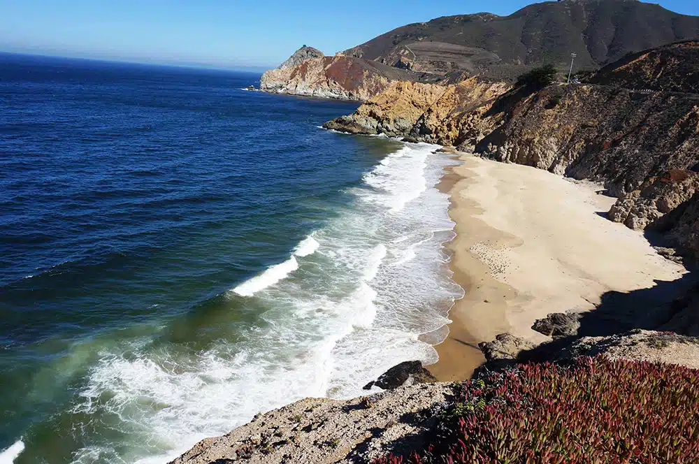 A high-angle, wide shot of a secluded sandy beach nestled against steep, rocky cliffs under a clear blue sky.