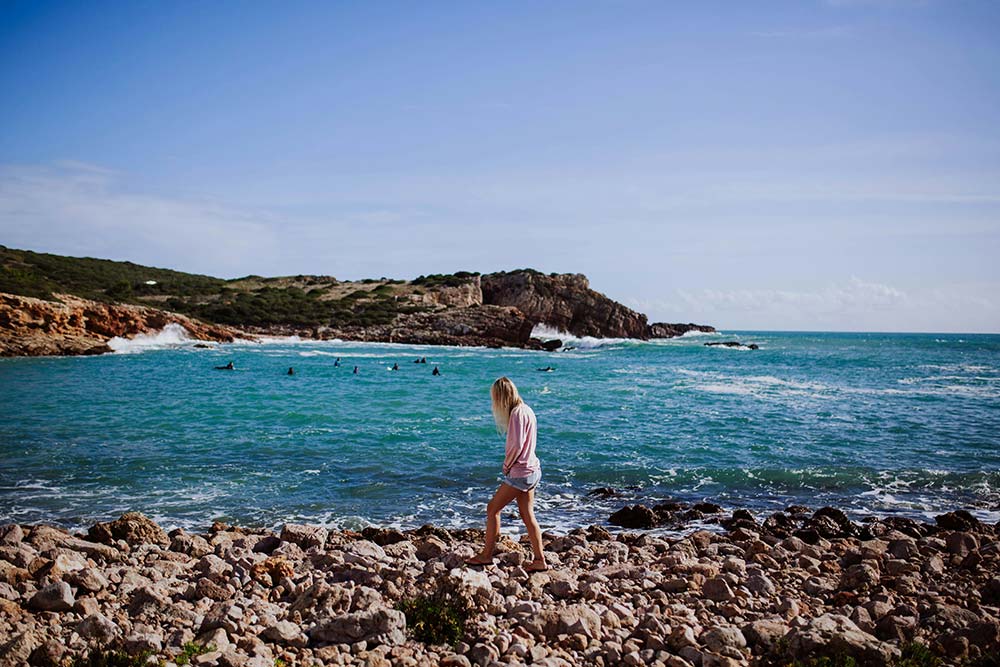 A lone woman with blonde hair walks across a rugged, rocky shoreline in the foreground