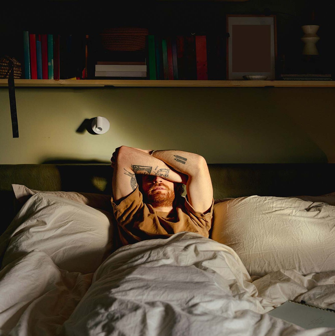 A man lying in bed, covering his face in low light, expressing distress, shame, and emotional withdrawal
