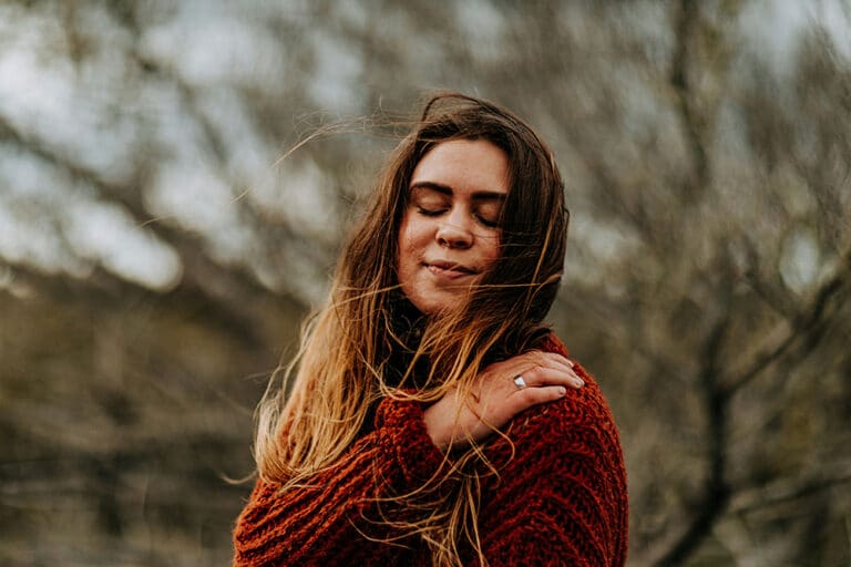 A woman with long, brown hair stands outdoors with her eyes closed and a peaceful expression.