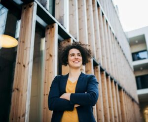 A woman standing with arms folded in front of a building, smiling confidently and expressing strength and self-assurance.
