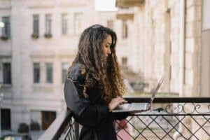 A woman standing on a balcony working on a laptop, reflecting independence, focus, and balancing personal growth.