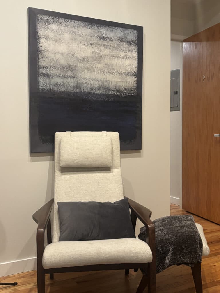Cream lounge chair with dark pillow and throw, under abstract gray-and-black wall art modern, calm corner beside wood door.