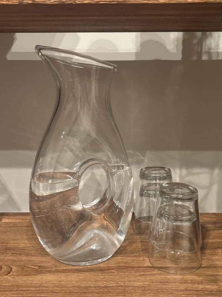 Clear glass water pitcher with handle, half-filled, beside three upside-down glasses on wood shelf simple, modern hydration setup.