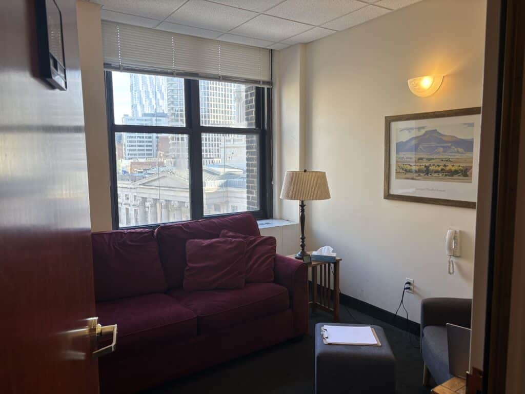 Maroon sofa by window with city view, beside lamp, side table, framed landscape art, and wall sconce cozy office lounge with urban backdrop.