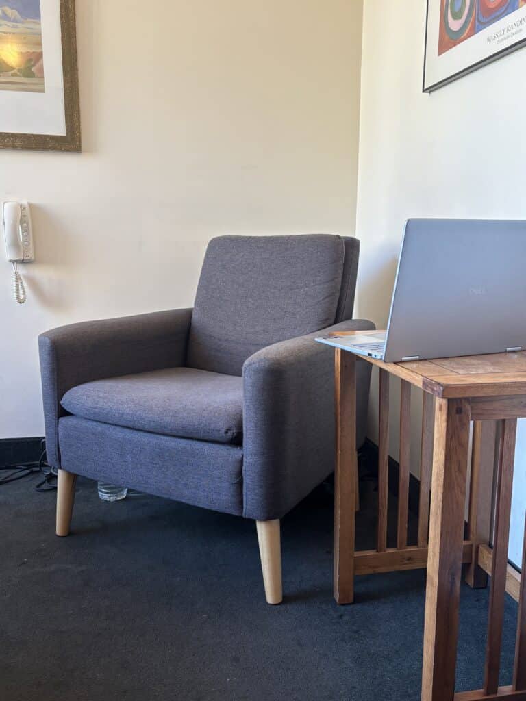 Gray armchair with light wood legs beside wooden table holding open laptop minimalist office nook with wall art and landline phone.