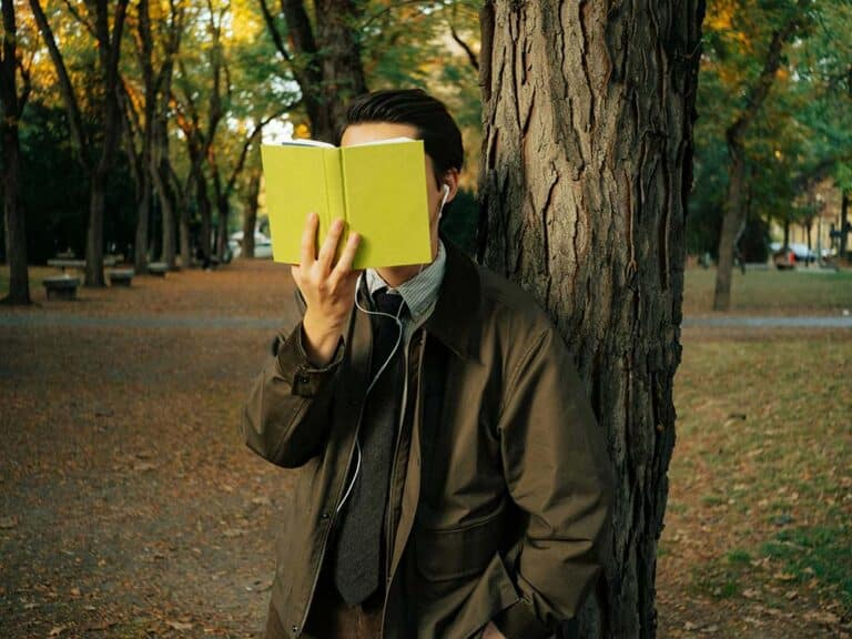 Man in suit reads book leaning against tree