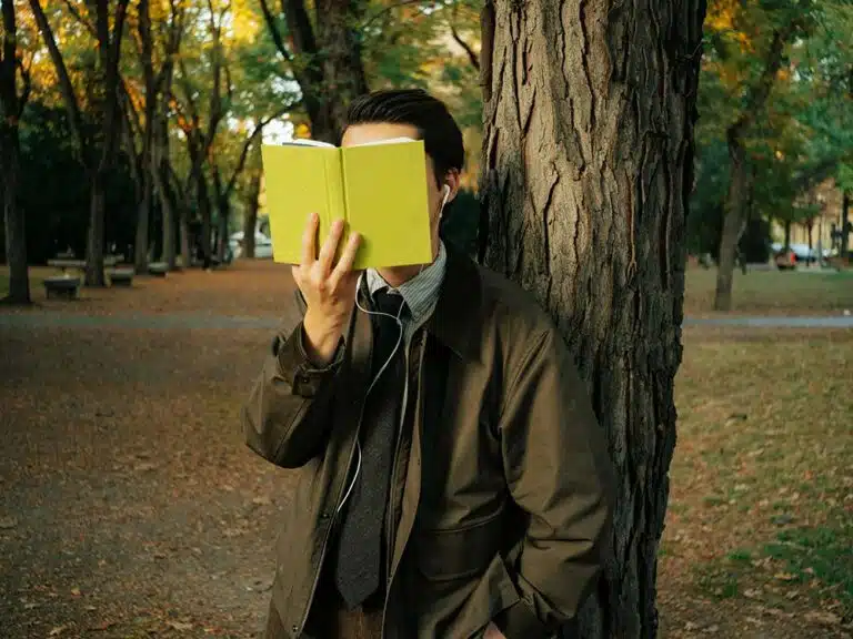 Man in suit reads book leaning against tree
