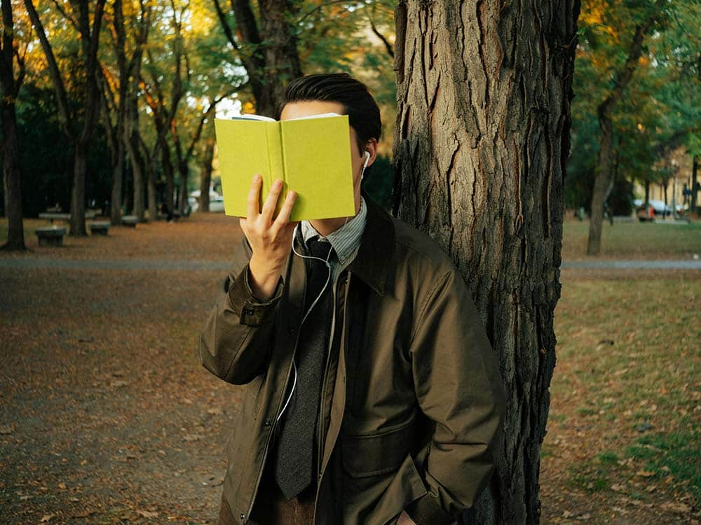 Man in suit reads book leaning against tree