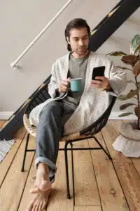 A young man enjoying a relaxed morning routine, checking his mobile phone with a hot drink in a modern apartment near a staircase